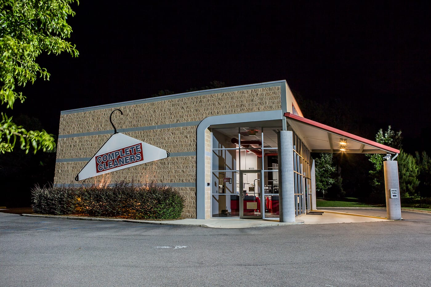 The Carrington store at night, side view with the Complete Cleaners hanger logo on the brick wall and the drive-thru lane to the right